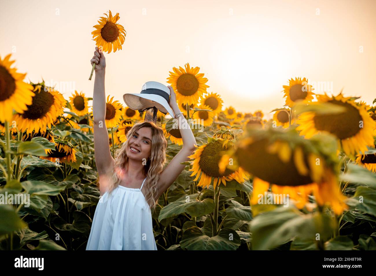 A woman in a white dress smiles while holding a sunflower and her hat ...