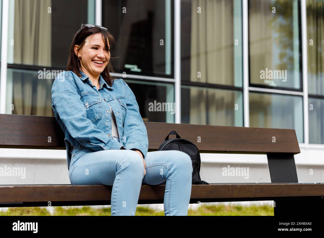 A woman wearing a denim jacket and blue jeans sits on a bench outside a ...
