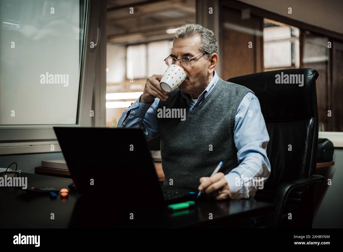 A man sits at a desk in an office, drinking coffee from a mug and working on a laptop. Stock Photo