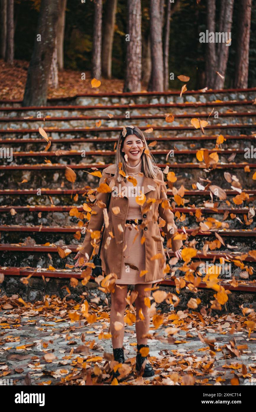 A woman in a brown coat stands in a forest, smiling as leaves fall around her. Stock Photo