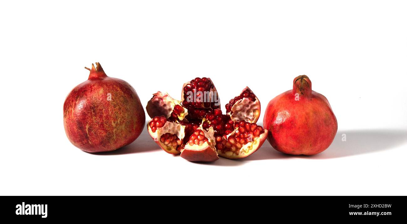 Three pomegranate fruits are depicted on a white background, one of which is broken into pieces. Stock Photo