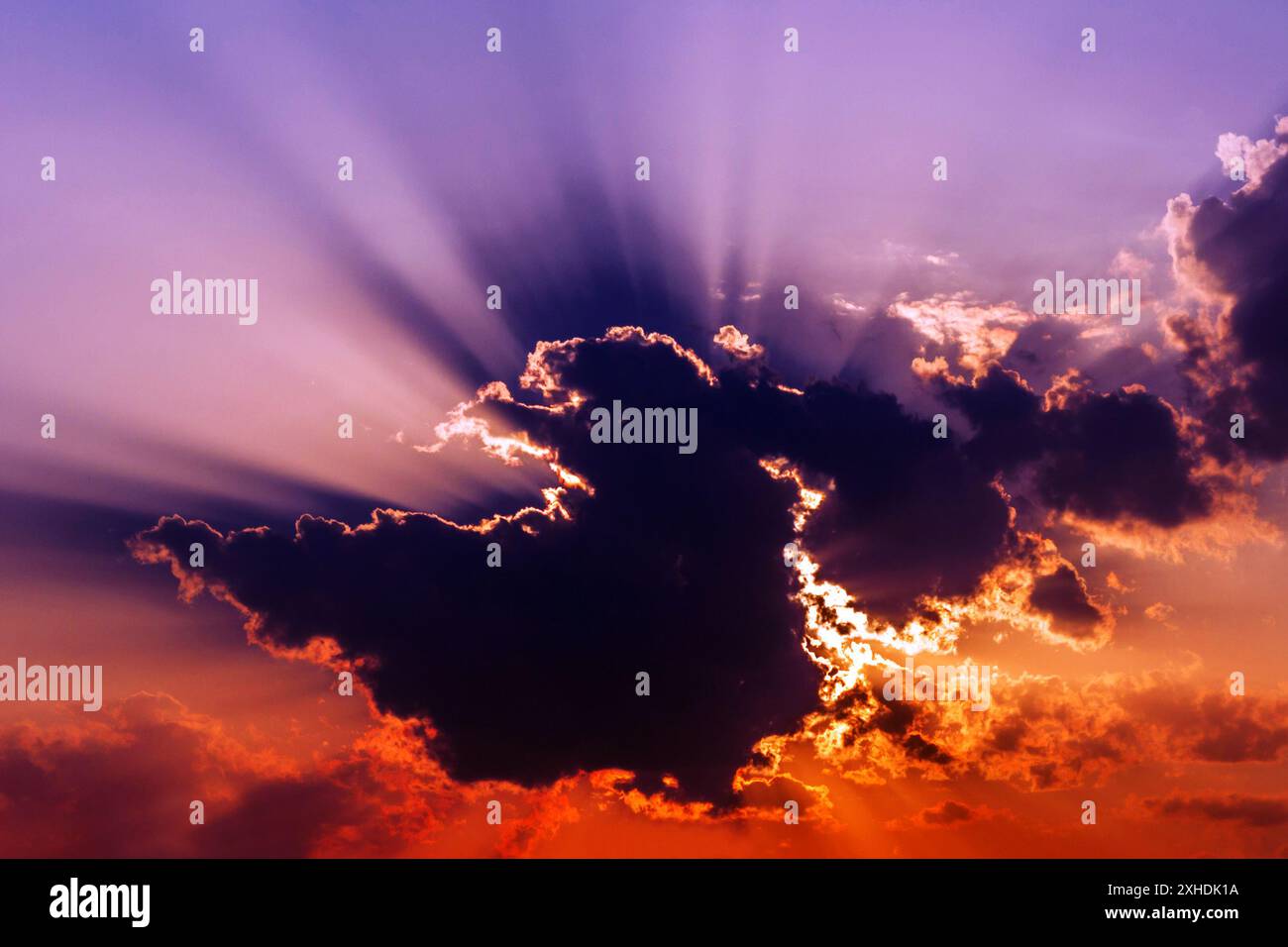 A vibrant sunset with golden rays of light piercing through a large, dark cloud, illuminating the surrounding clouds in a fiery orange glow. Stock Photo