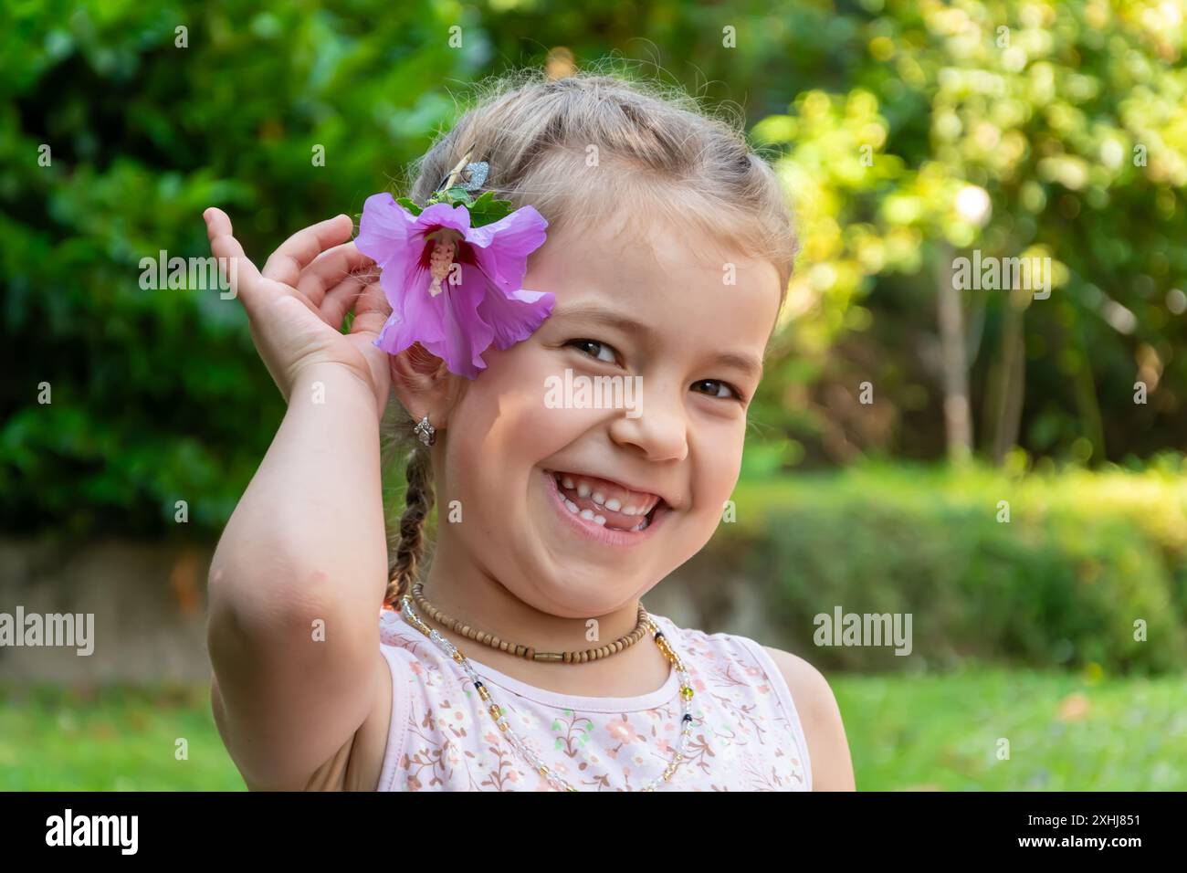 Portrait of a laughing 6 year old girl with a flower in her hair against the background of a blooming garden. The girl's milk teeth have fallen out. Stock Photo