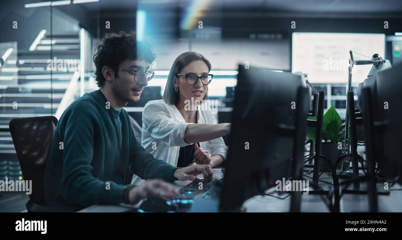 Two Young Colleagues Working on Computers and Talking at a Workplace. Female and Male Software Developers Discussing a Solution for Their Collaborative Artificial Intelligence Project Stock Photo