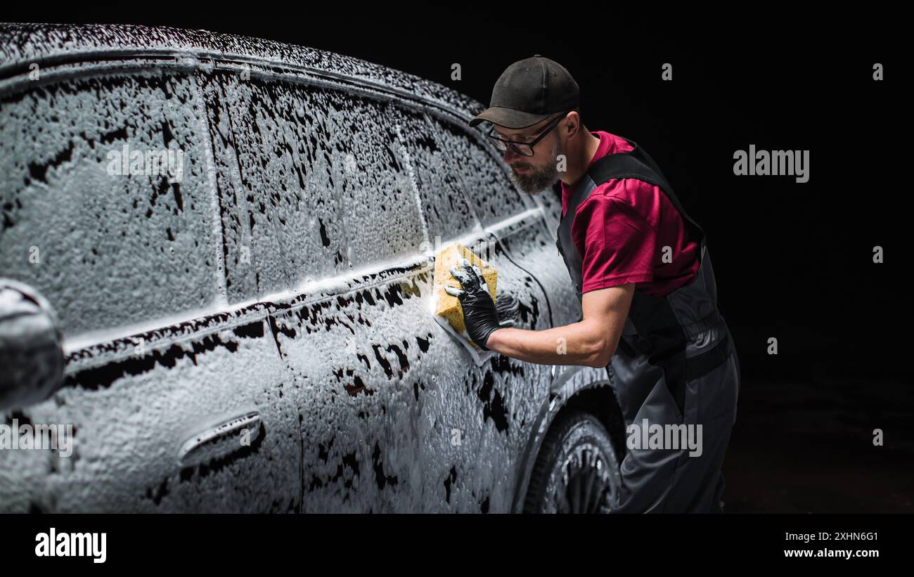 Portrait of an Adult Man Working in a Detailing Studio, Prepping a Factory Fresh Electric SUV for Maintenance Work and Car Care Treatment. Cleaning Technician Using Sponge with Soap Water Stock Photo