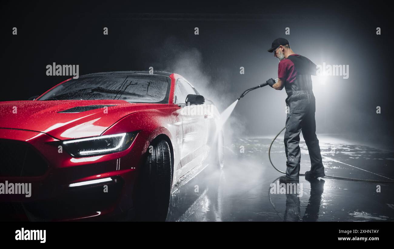 Portrait of an Adult Man Working in a Detailing Studio, Prepping a Factory Fresh Red Sportscar for Maintenance Work and Car Care Treatment. Cleaning Technician Using High Pressure Washer Stock Photo