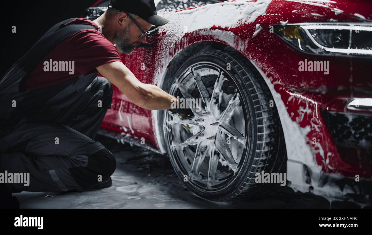 Car Ad Style Photo of a Professional Car Wash Specialist Using a Big Soft Sponge to Wash the Rims of a Beautiful Red Sportscar with Shampoo Before Detailing, Polishing and Waxing Stock Photo