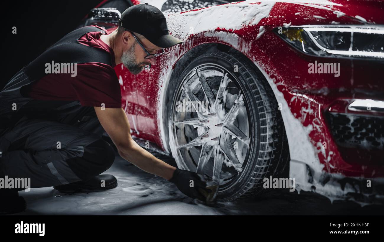 Car Ad Style Photo of a Professional Car Wash Specialist Using a Big Soft Sponge to Wash the Rims of a Beautiful Red Sportscar with Shampoo Before Detailing, Polishing and Waxing Stock Photo