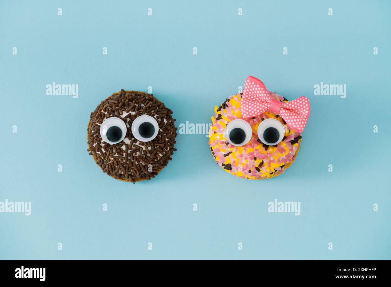 Two sweet cookies with colorful sprinkles with googly eyes and one with pink hair bow on light pastel blue background. Funny sweet food concept. Stock Photo