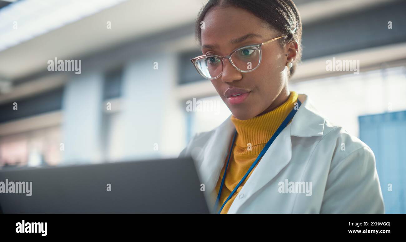 Portrait of Young Black Female Expert in a Lab Coat Smiling and Using Laptop Computer. Professional and Successful Woman Working as an Engineer, Developing High Technology Projects Stock Photo