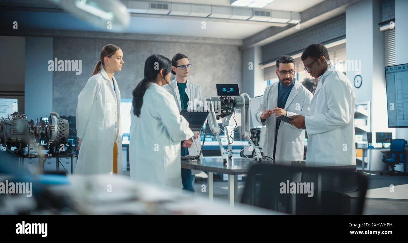 Diverse Team of Industrial Robotics Specialists Gathered Around a Table With a Mobile Robot. Engineers In Lab Coats Discussing an Automated AI Robotic Dog, Using Laptop and Tablet, Brainstorming Stock Photo