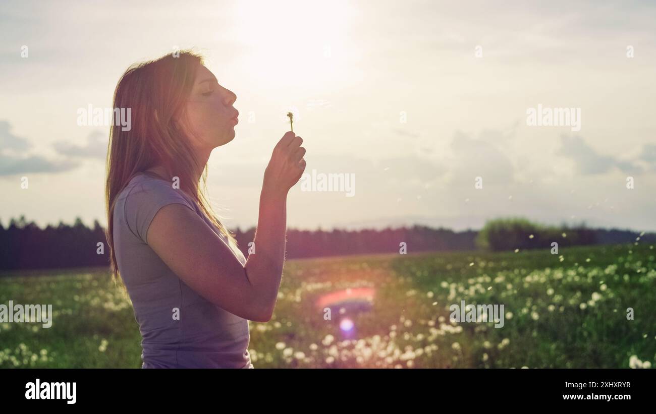 Portrait of a beautiful young woman blowing a dandelion in a flower meadow. Femininity and freedom concepts. Stock Photo