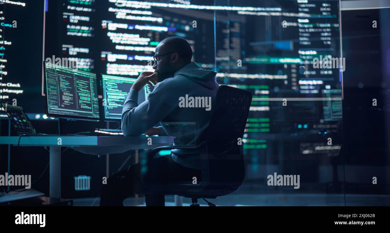 Portrait of a Black Man Working on Computer, Typing Lines of Code that Appear on Big Screens Surrounding him in Monitoring Room. Male Programmer Creating Innovative Software Using AI Data and System Stock Photo