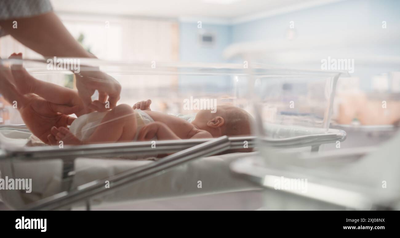 Attentive Mother Changing Diaper to a Newborn Baby. Neonate Child Lying in a Hospital Crib in a Nursery Clinic Facility. Motherhood, Childhood and Medical Concept Stock Photo