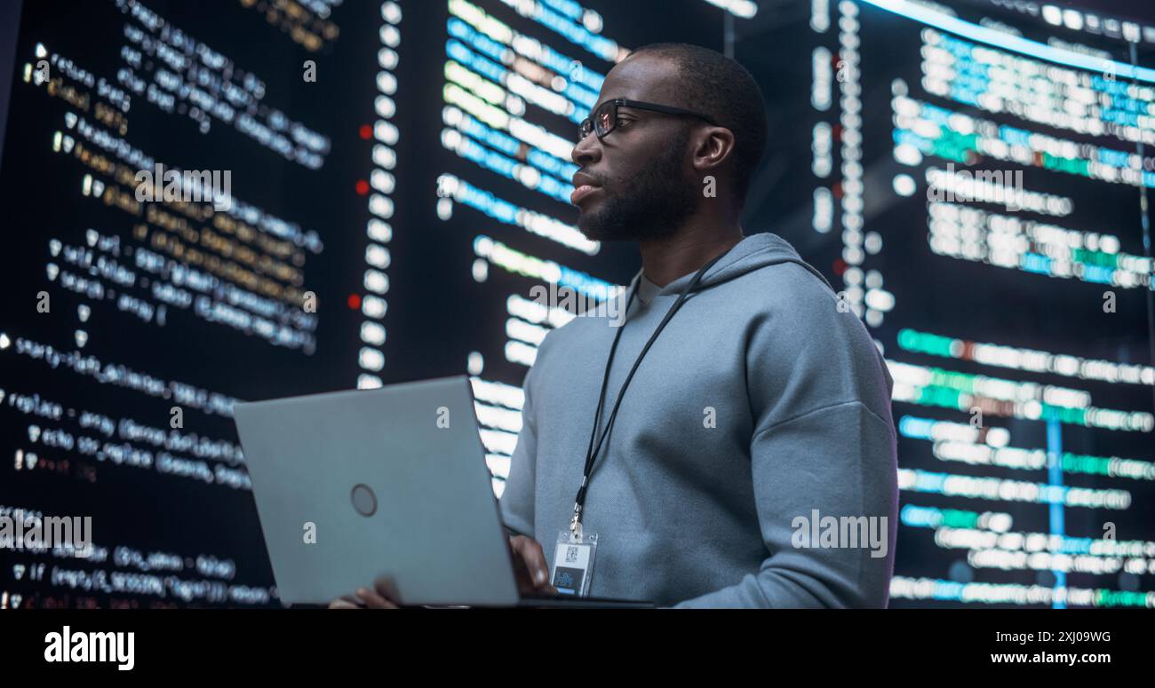 Portrait of Young Black Man Working on Laptop Computer, Looking at Big Digital Screen Displaying Back-end Code Lines. Professional Programmer Developing a Big Data Interface Software Project Concept. Stock Photo