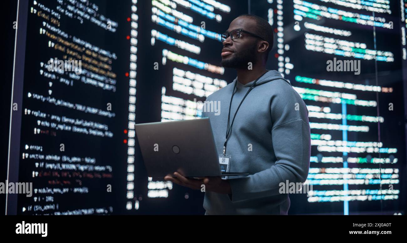 Portrait of Young Black Man Working on Laptop Computer, Looking at Big Digital Screen Displaying Back-end Code Lines. Professional Programmer Developing Big Data Interface Software Project. Stock Photo