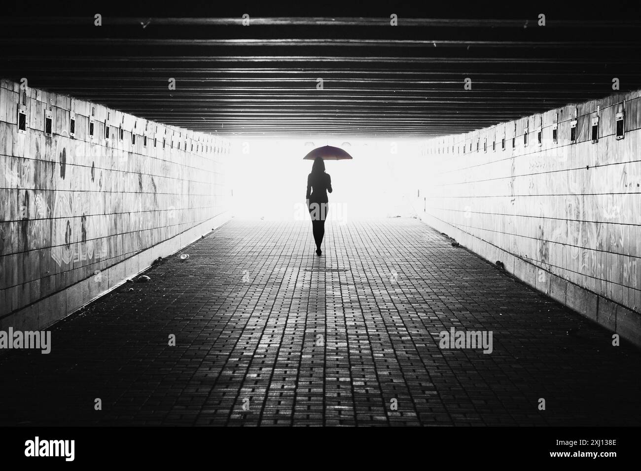 Silhouette of woman with umbrella in a tunnel. Black and white image with light coming from behind. Person walking away from the camera. Stock Photo