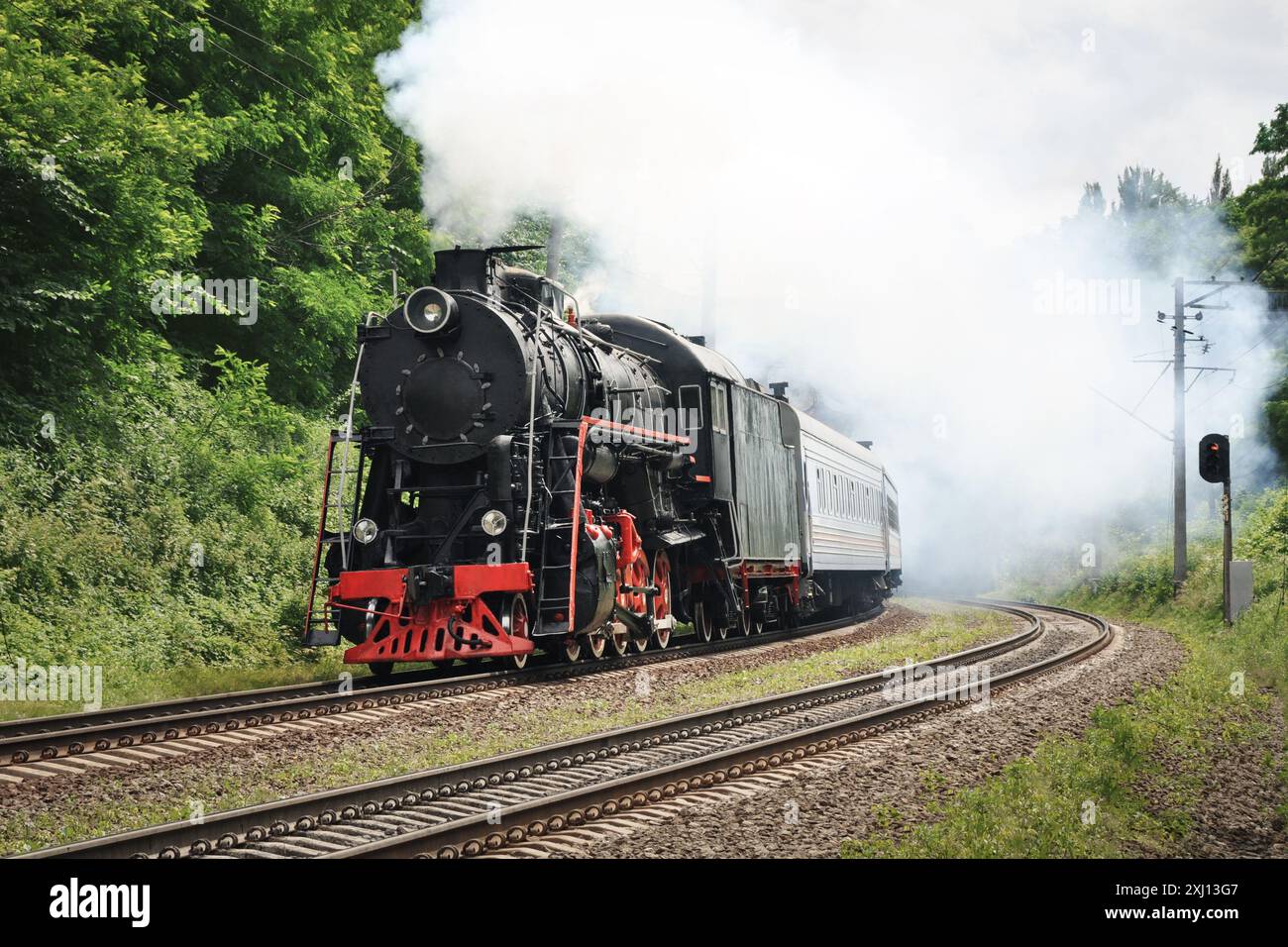 Old steam engine locomotive driving by in a green forest. Black vintage locomotive takes a curve while pulling a train on a sunny day. Stock Photo