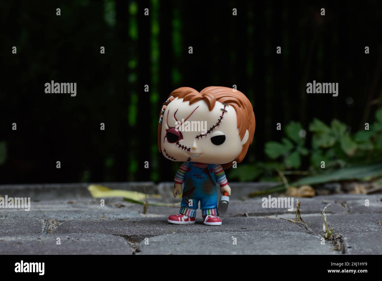 Funko Pop action figure of Chucky serial killer doll from popular horror movies Child's Play. Dark yard, black fence, shadows, moody green background. Stock Photo