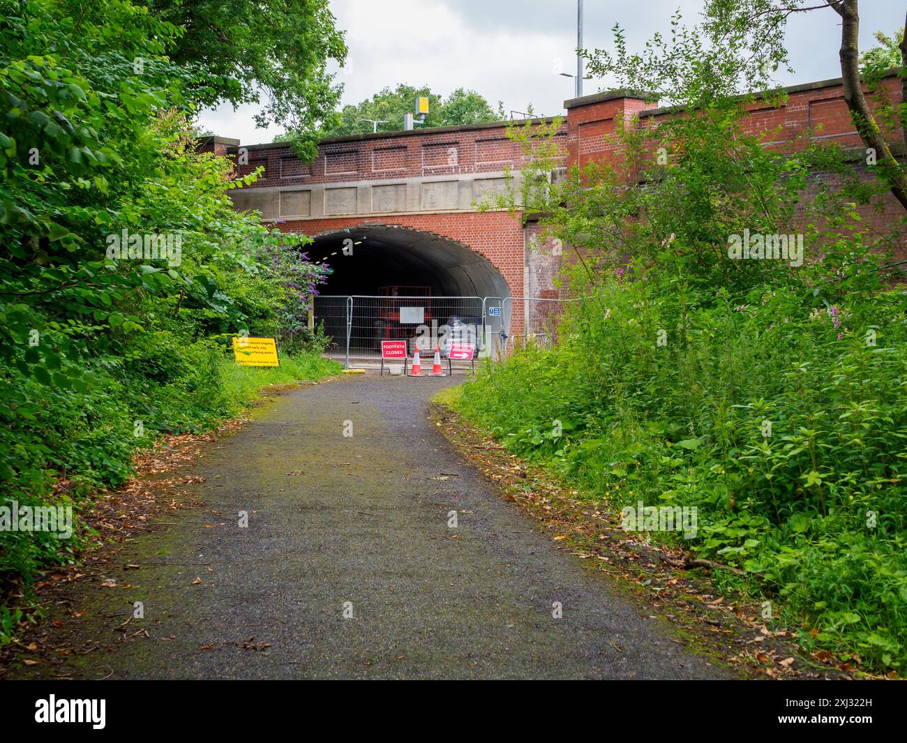 Roe Green loopline Worsely. Fenced-off tunnel entrance on a paved path surrounded by lush greenery and bright construction signs. Stock Photo