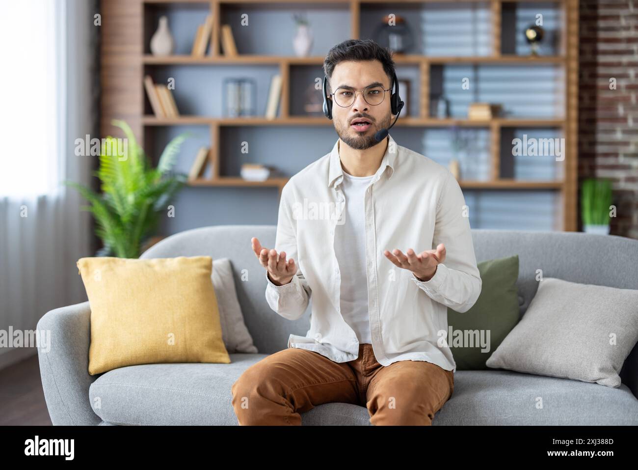 Worried young Indian man is at home sitting on the sofa in a headset talking nonsense, explaining, discussing to the camera, waving his arms. Stock Photo
