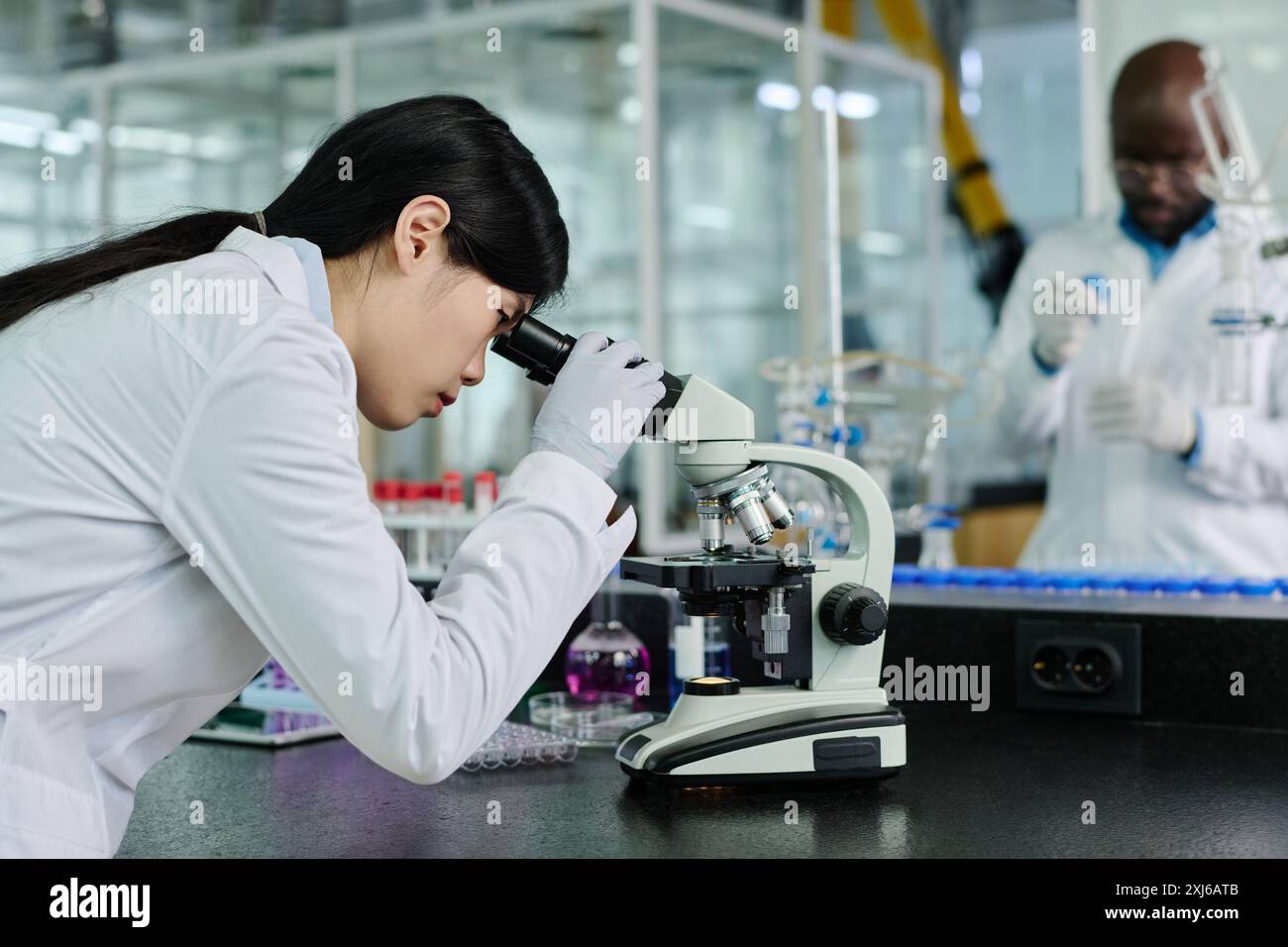 Side view of young Chinese female biochemist in whitecoat looking in microscope while studying new virus in chemical laboratory Stock Photo