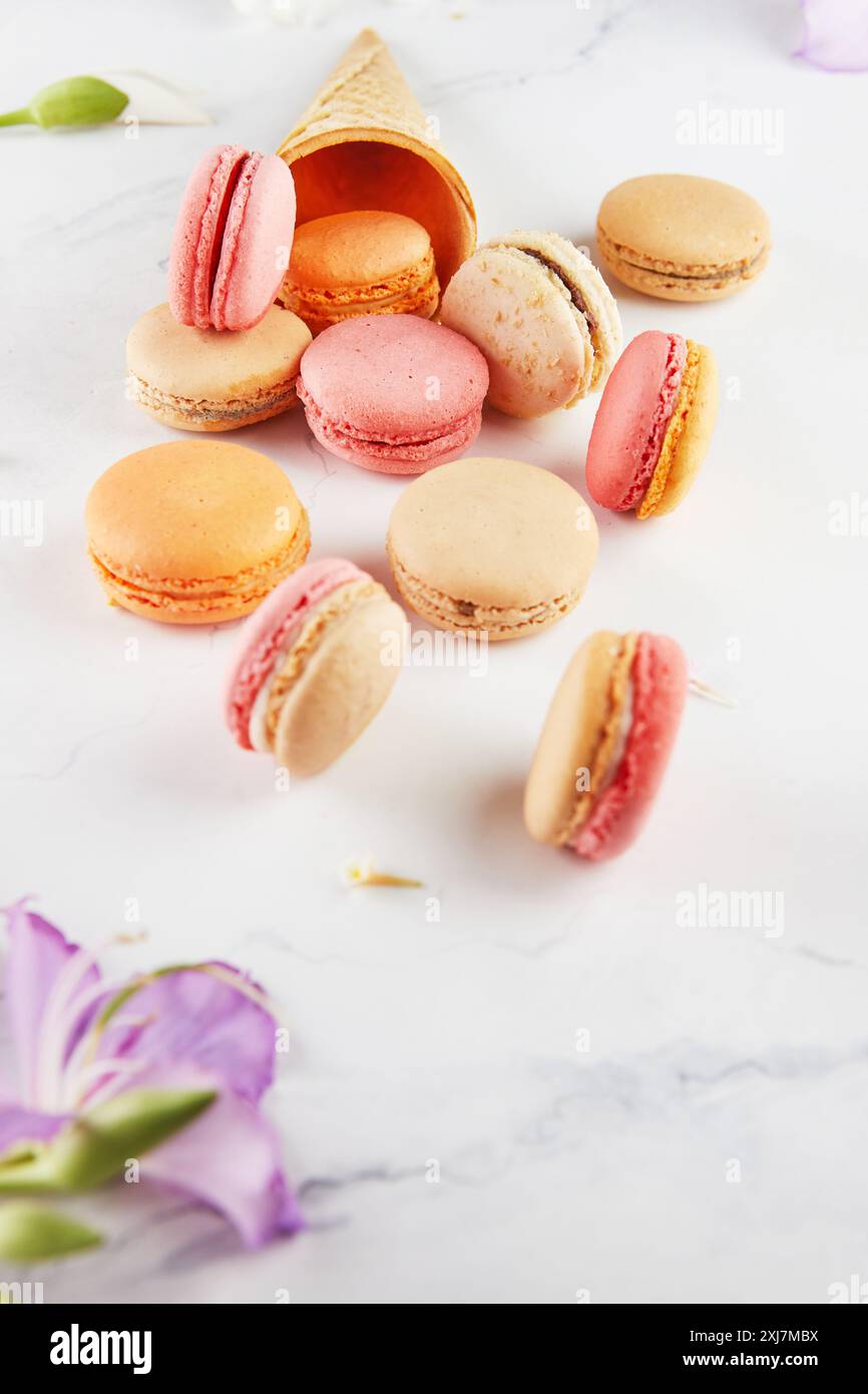 Assorted colorful macarons scattered with an empty ice cream cone and flowers on a white marble background. Stock Photo