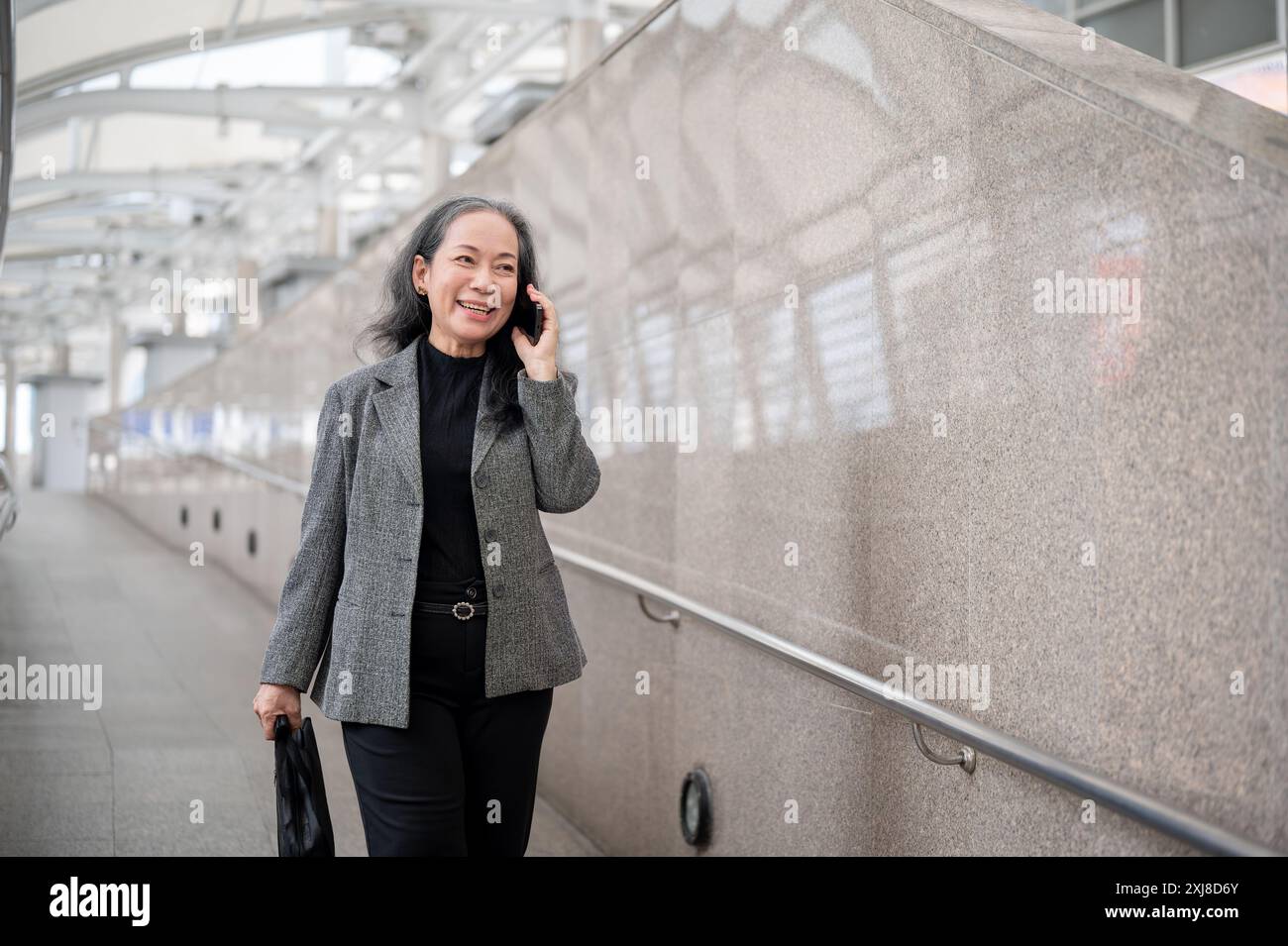 A successful, mature Asian businesswoman is talking on the phone with her client while walking on the skywalk or skytrain station, commuting to work i Stock Photo