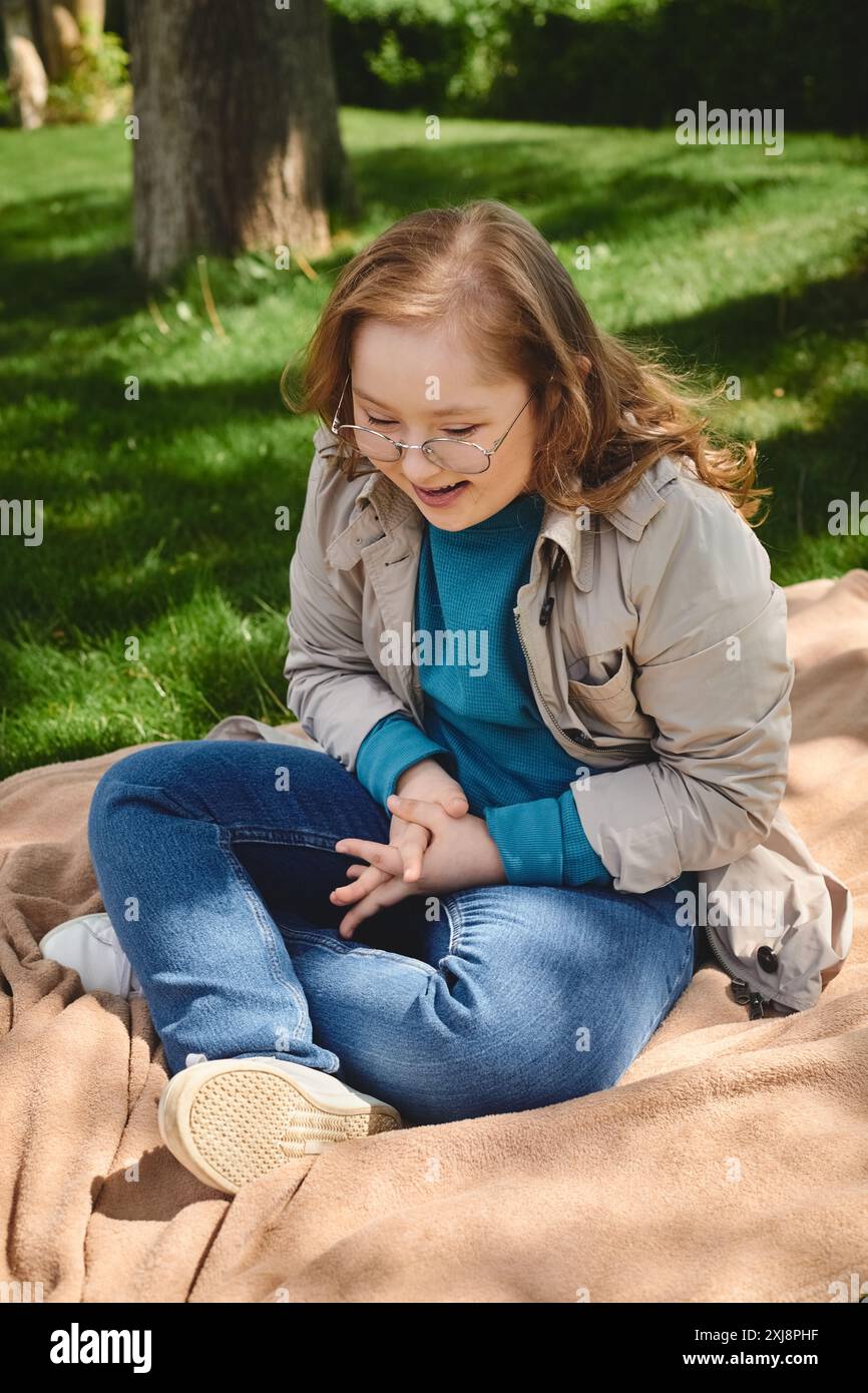 A little girl with Down syndrome sits on a blanket in a park, laughing. Stock Photo