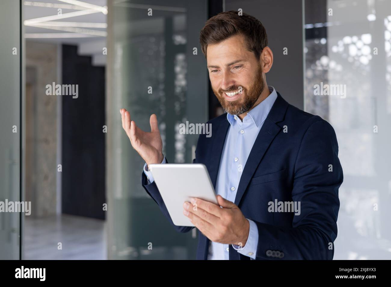 Confident, middle-aged businessman in a suit smiles while using a digital tablet in a stylish, contemporary office environment. Communicating or presenting with a touch of elegance. Stock Photo