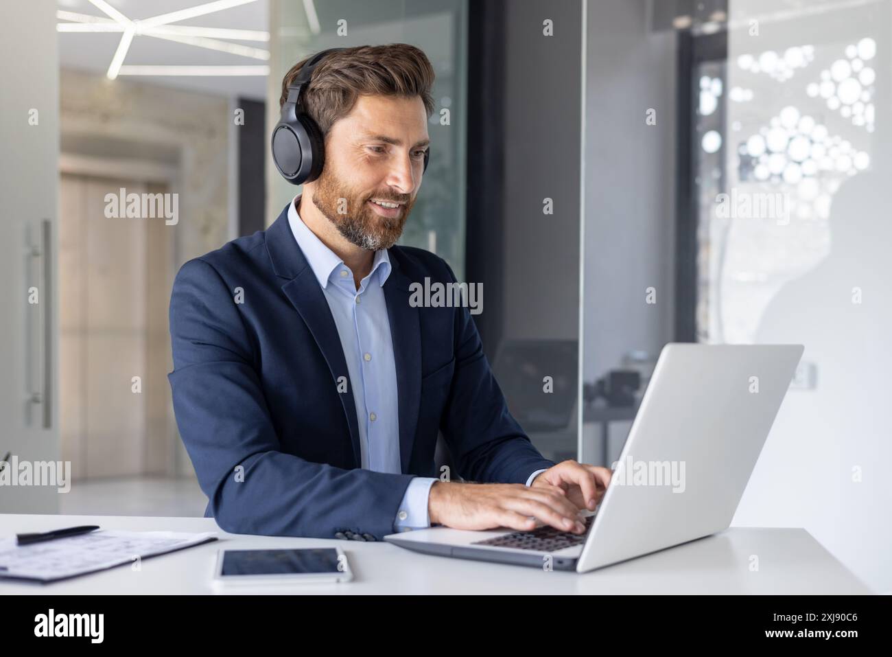 A cheerful businessman wearing headphones sits at a modern office desk working on a laptop, portraying focus and professionalism in a corporate setting. Stock Photo