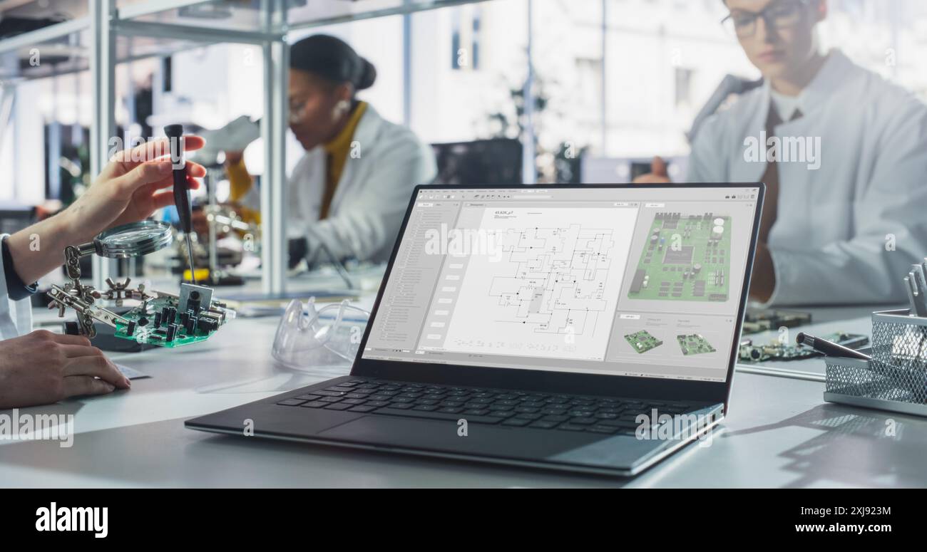 Laptop Computer with Technology Program Display Standing on a Table in a Science Class Laboratory. Diverse Students Practicing Soldering Electronic Components for Their Tech Projects Stock Photo