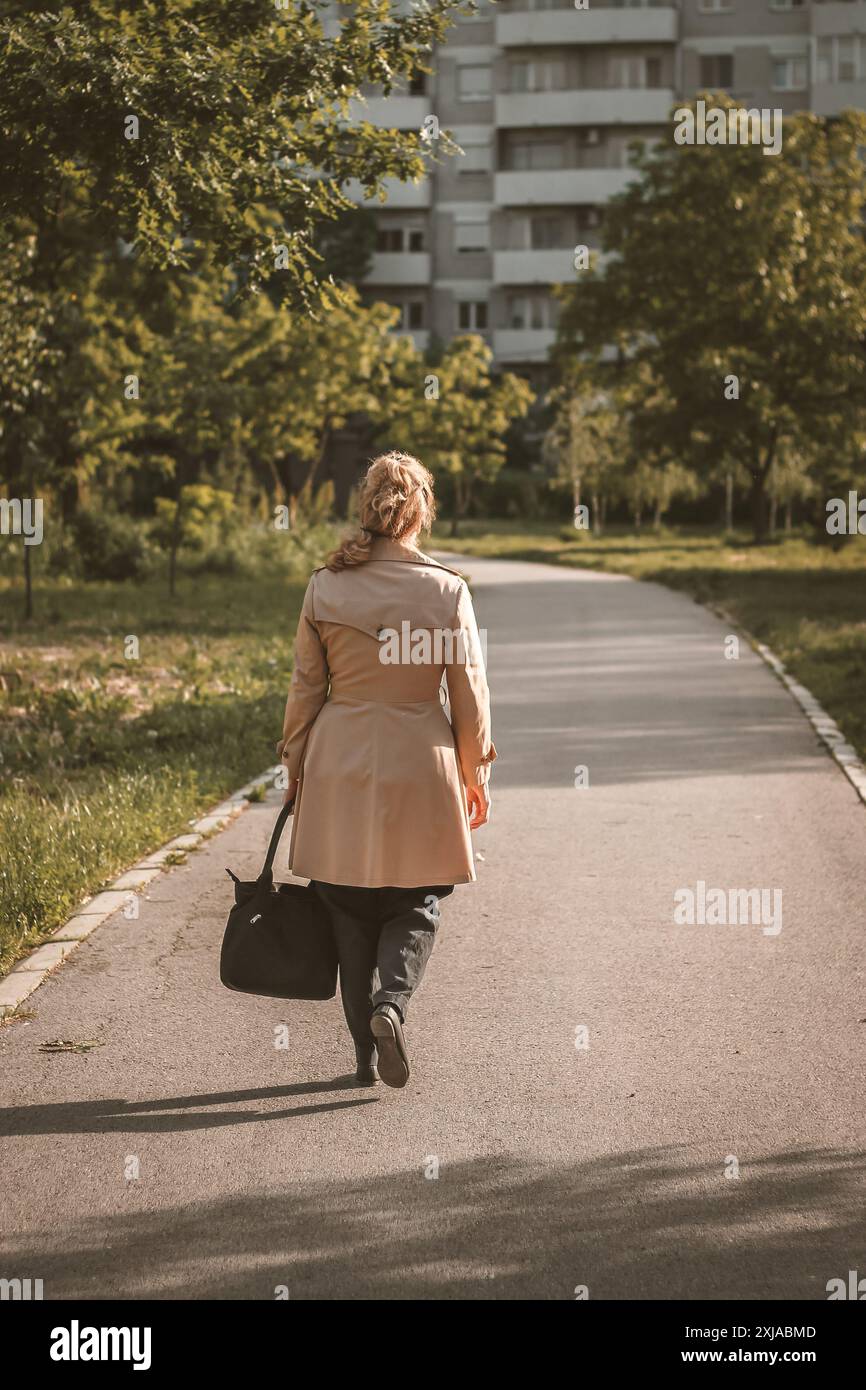 A woman in a beige coat walks down a paved pathway in a park. She ...