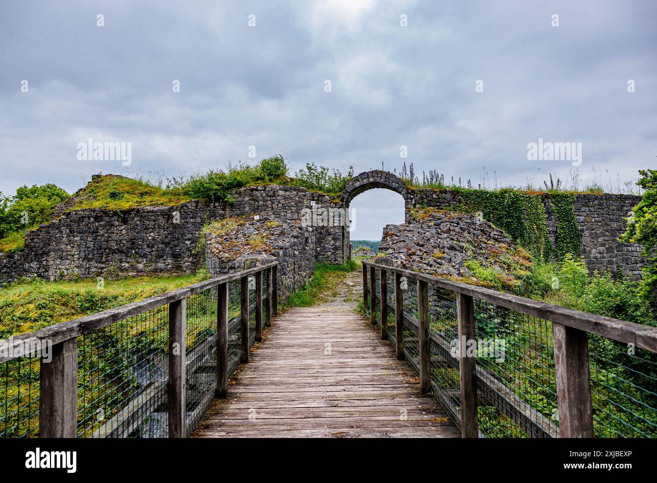 Wooden bridge leading to an arched gate at Logne Castle, time-worn stone wall against gray cloud-covered sky, cloudy day in Ferrieres, Belgium Stock Photo