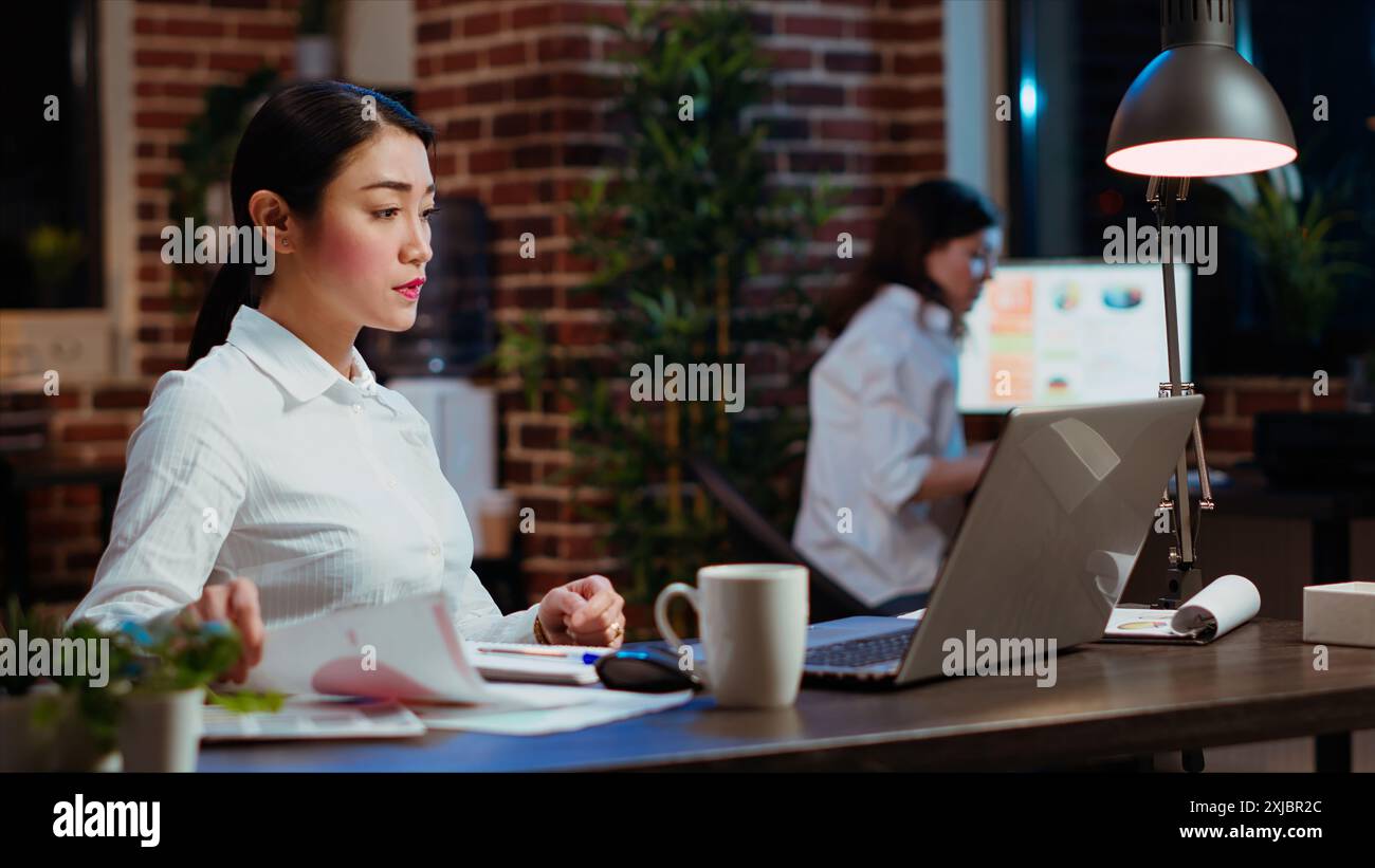 Asian bookkeeper working in office overnight, bored of checking financial data graphs and charts. Businesswoman trying to finish tedious paperwork, feeling stressed and extenuated Stock Photo