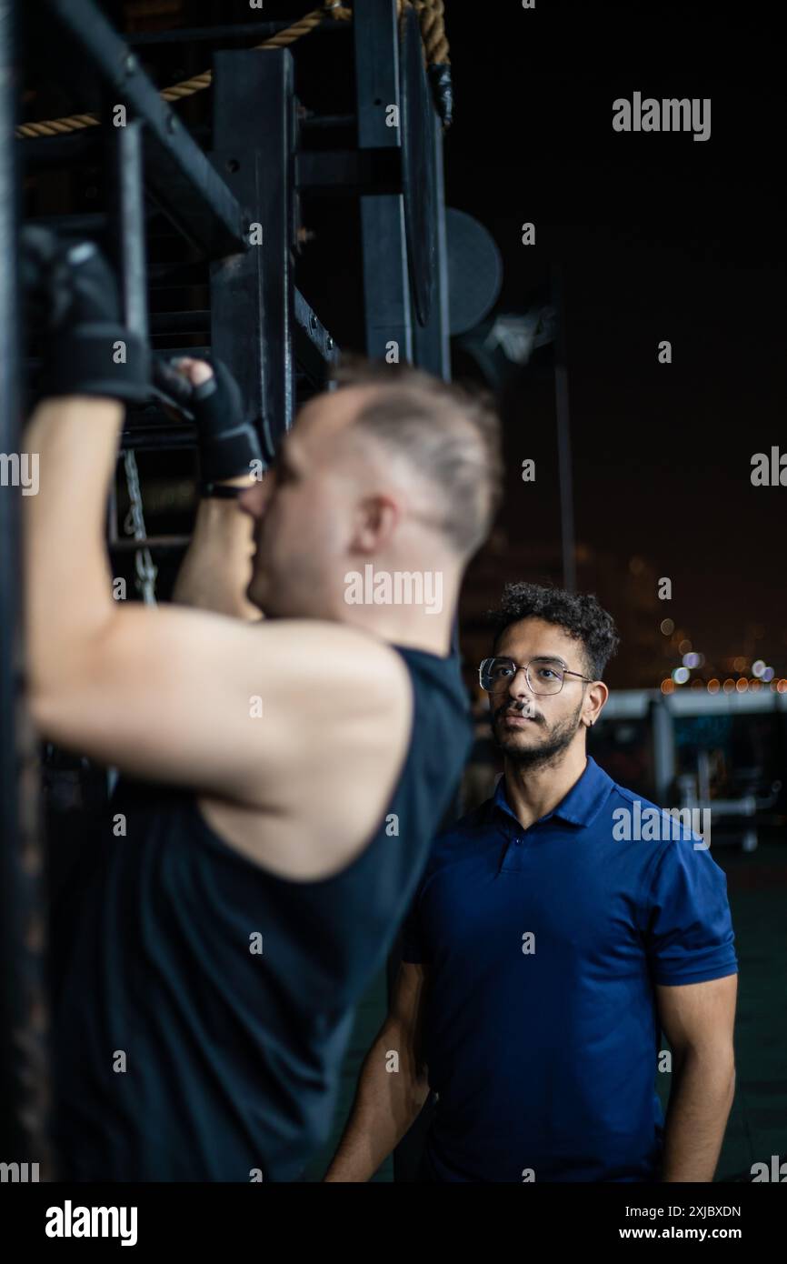 A man is doing pull-ups on a rig, while his trainer watches. Stock Photo