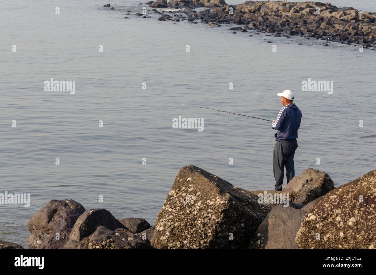 Man using a fishing rod to catch fish from the sea standing on rocks, Mumbai, Maharashtra Stock Photo
