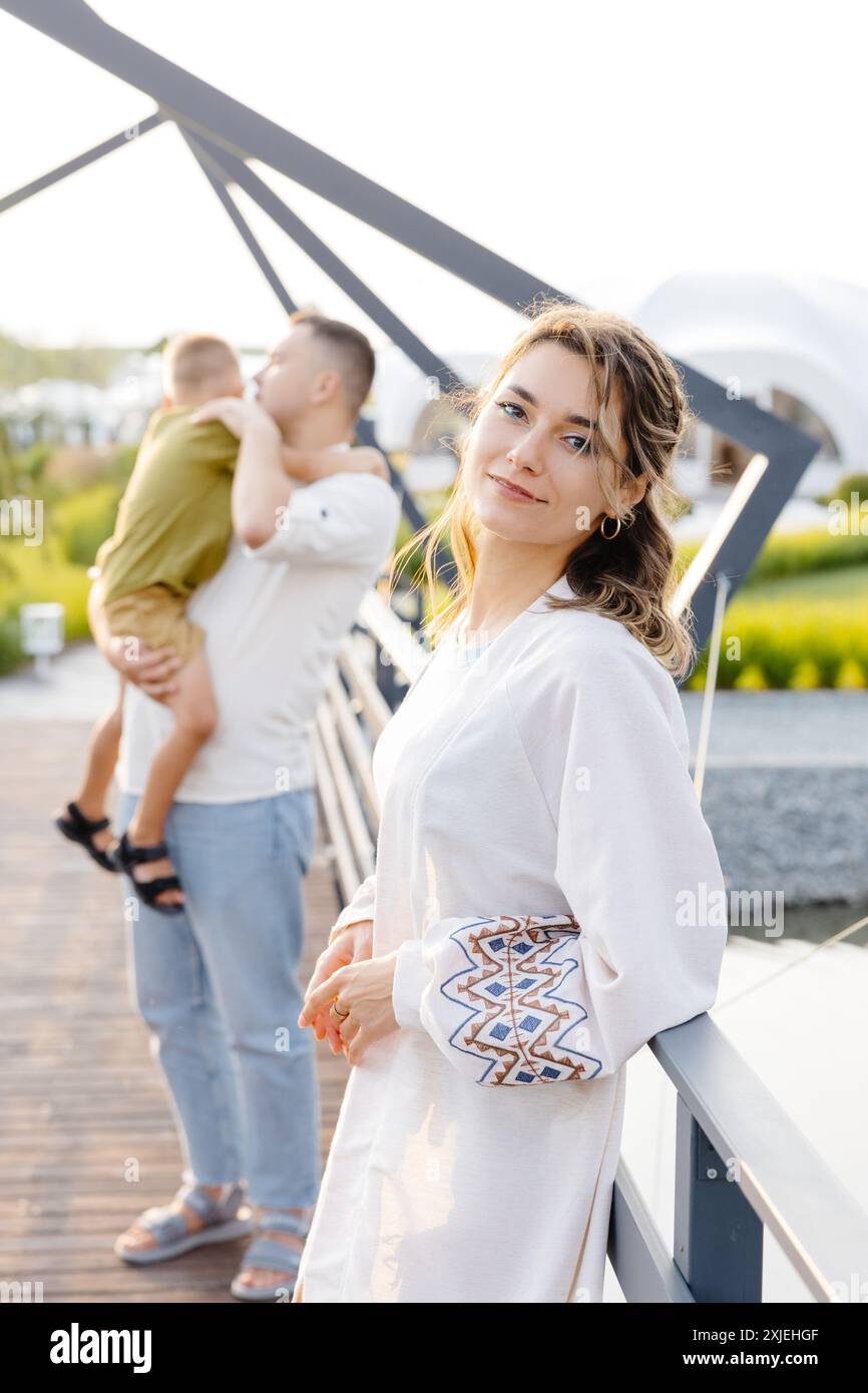 A woman in a white dress stands on a bridge, leaning against a railing and looking at the camera. Her husband holds their young son in his arms in the Stock Photo