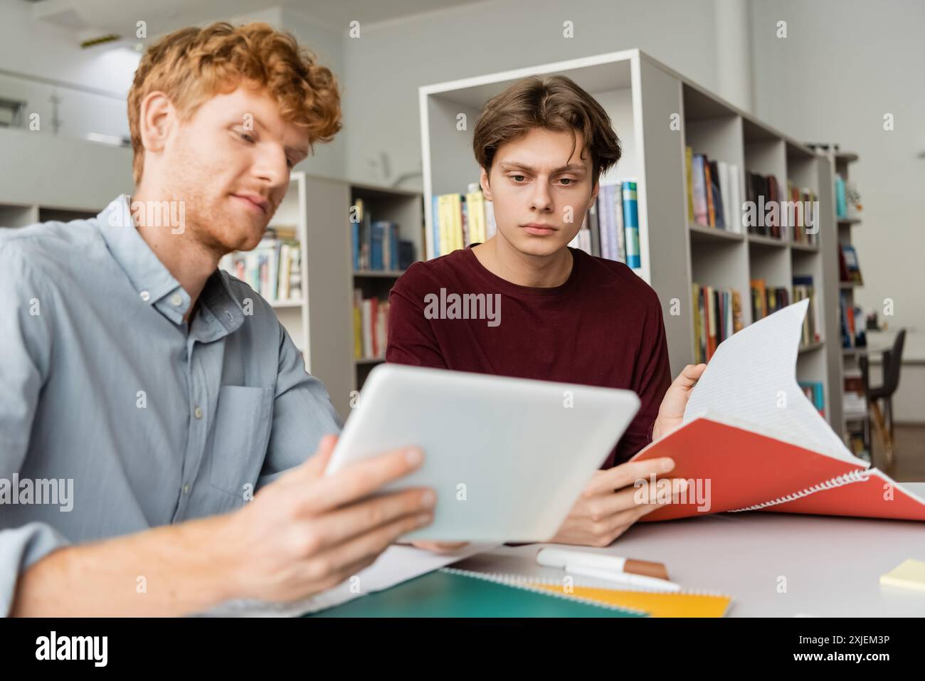 Two students engage with a tablet while seated at a table in a library. Stock Photo