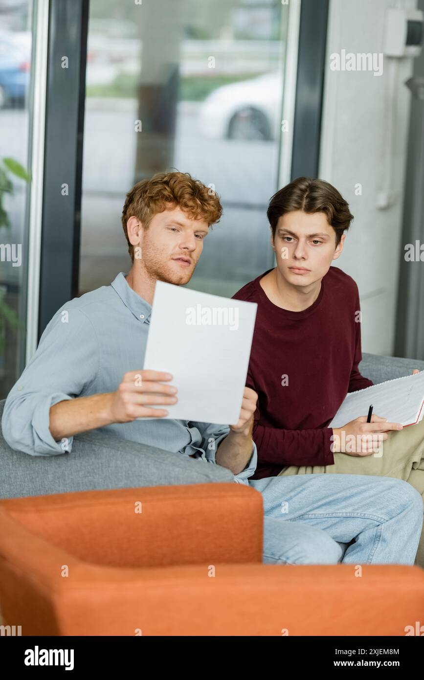 Two male students deeply engaged in studying papers on couch. Stock Photo