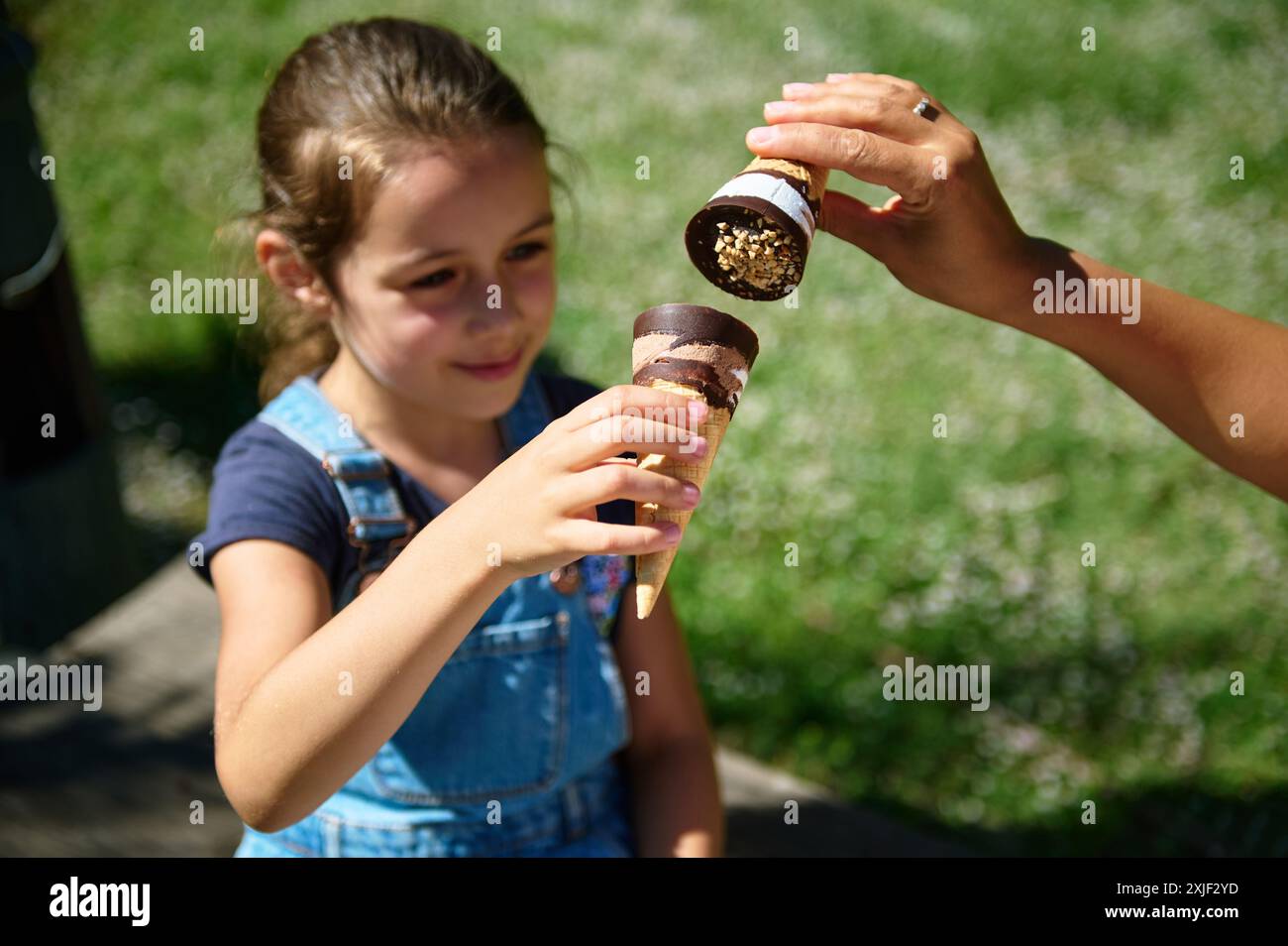 A young girl in a blue dress enjoys an ice cream cone handed by an adult on a sunny day. The background is green with a natural outdoor setting. Stock Photo