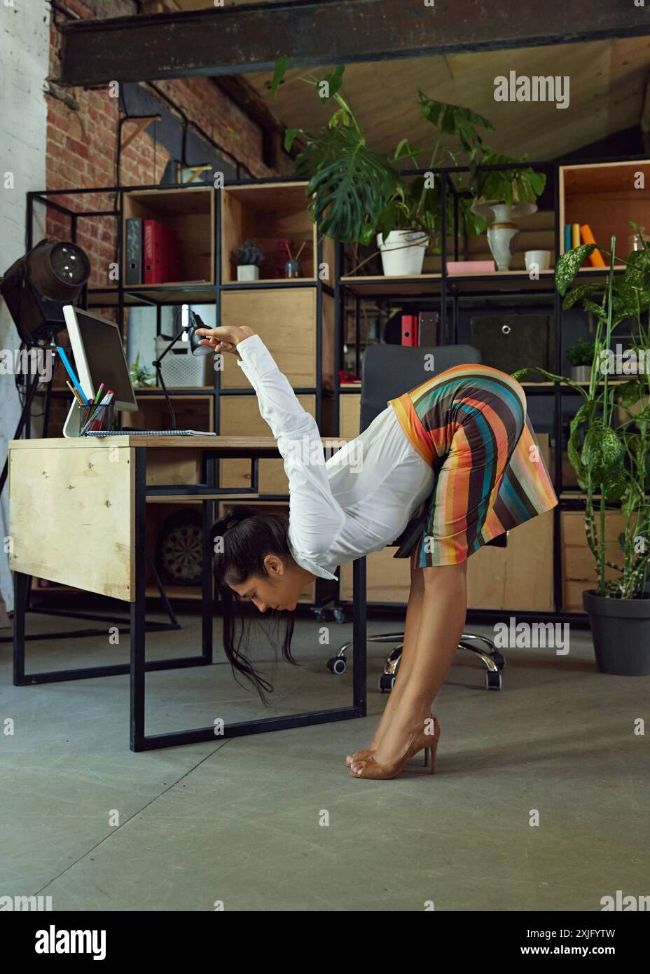 Woman, office employee in formal wear doing stretching at her desk while working, finishing tasks. Modern office setting. Comfort Stock Photo