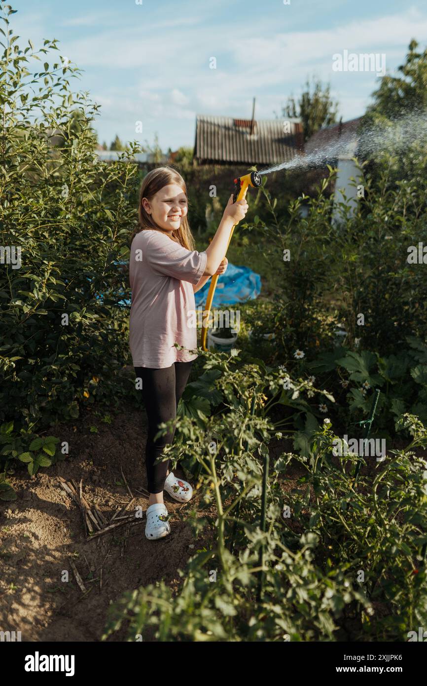 A girl is watering plants in garden with hose on sunny summer day Stock Photo