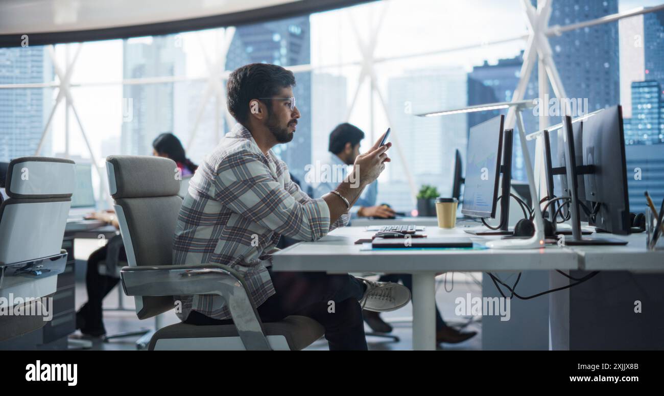Portrait of a Handsome Indian Manager Using Smartphone While Sitting Behind a Desk in a Corporate Office with a Group of Colleagues. Smiling Happy Man Answering a Message, Writing a Funny Online Post Stock Photo