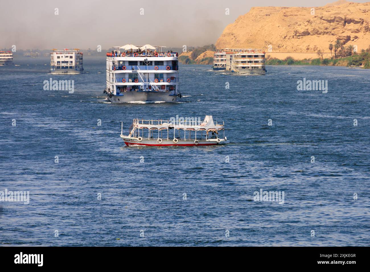 Local passenger ferry crosses the River Nile while avoiding large tourist cruise ships in convoy. Smoke haze and smog is visable from the ships exhaus Stock Photo