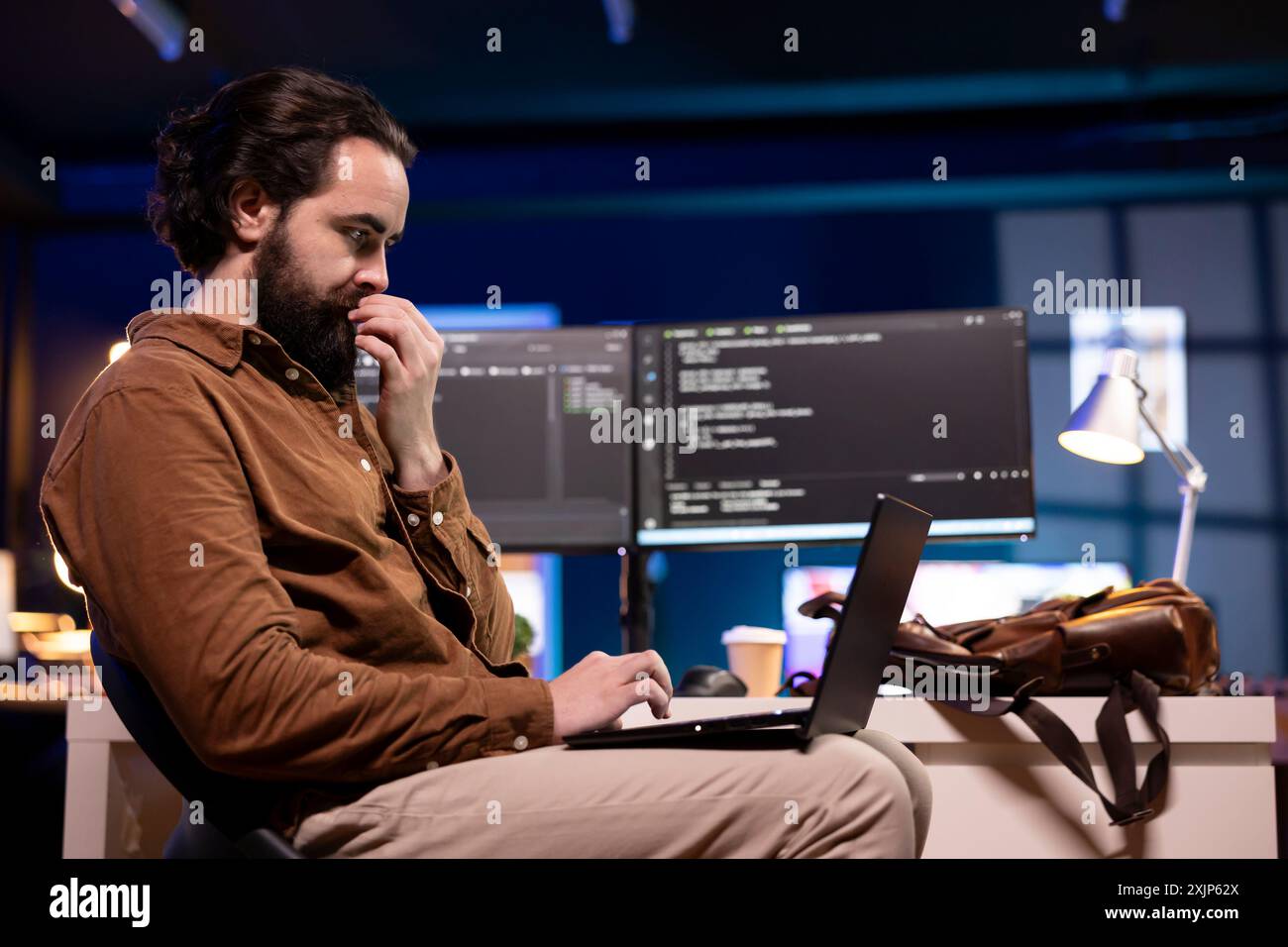 IT specialist working from home using laptop, testing and deploying programs and systems. Programmer checking code displayed on computer monitors, sitting at desk chair in apartment Stock Photo