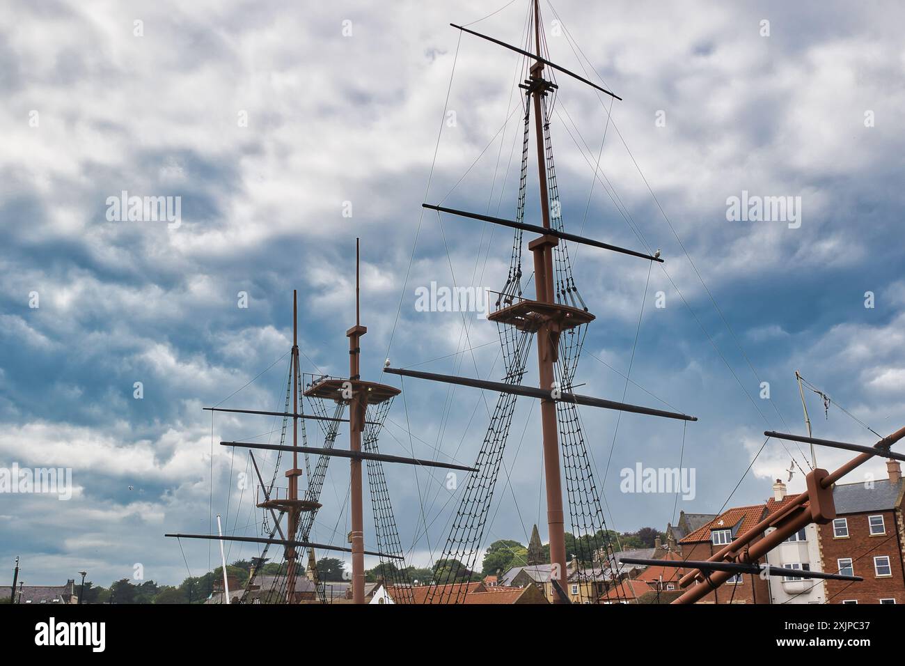 Three tall ship masts with rigging against a cloudy sky, with rooftops ...