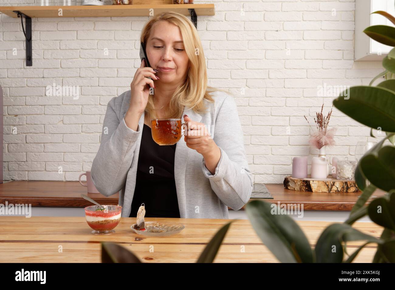 A middle-aged woman enjoying a phone chat in her cozy kitchen. She is sipping tea and eating dessert, creating a blissful moment. The inviting atmosph Stock Photo