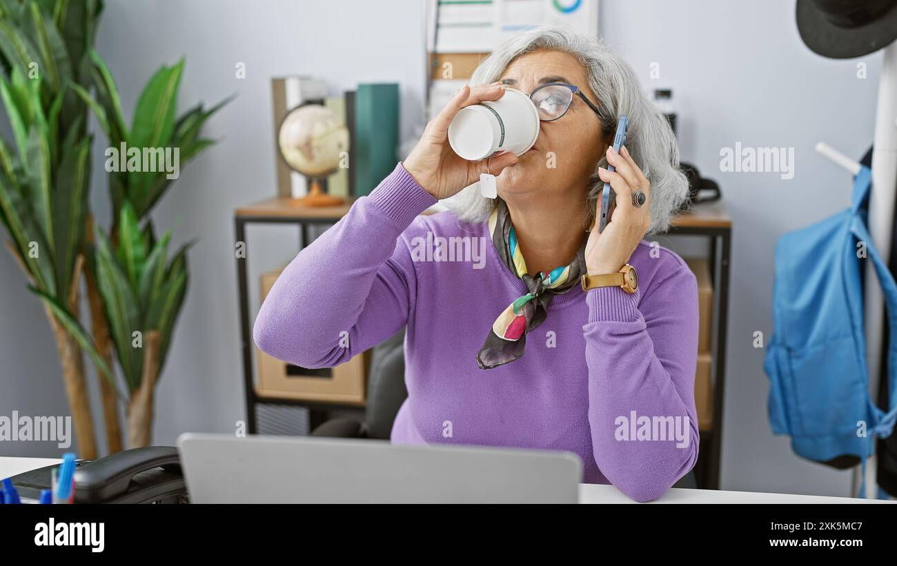 A mature woman multitasks in an office, drinking coffee and talking on ...