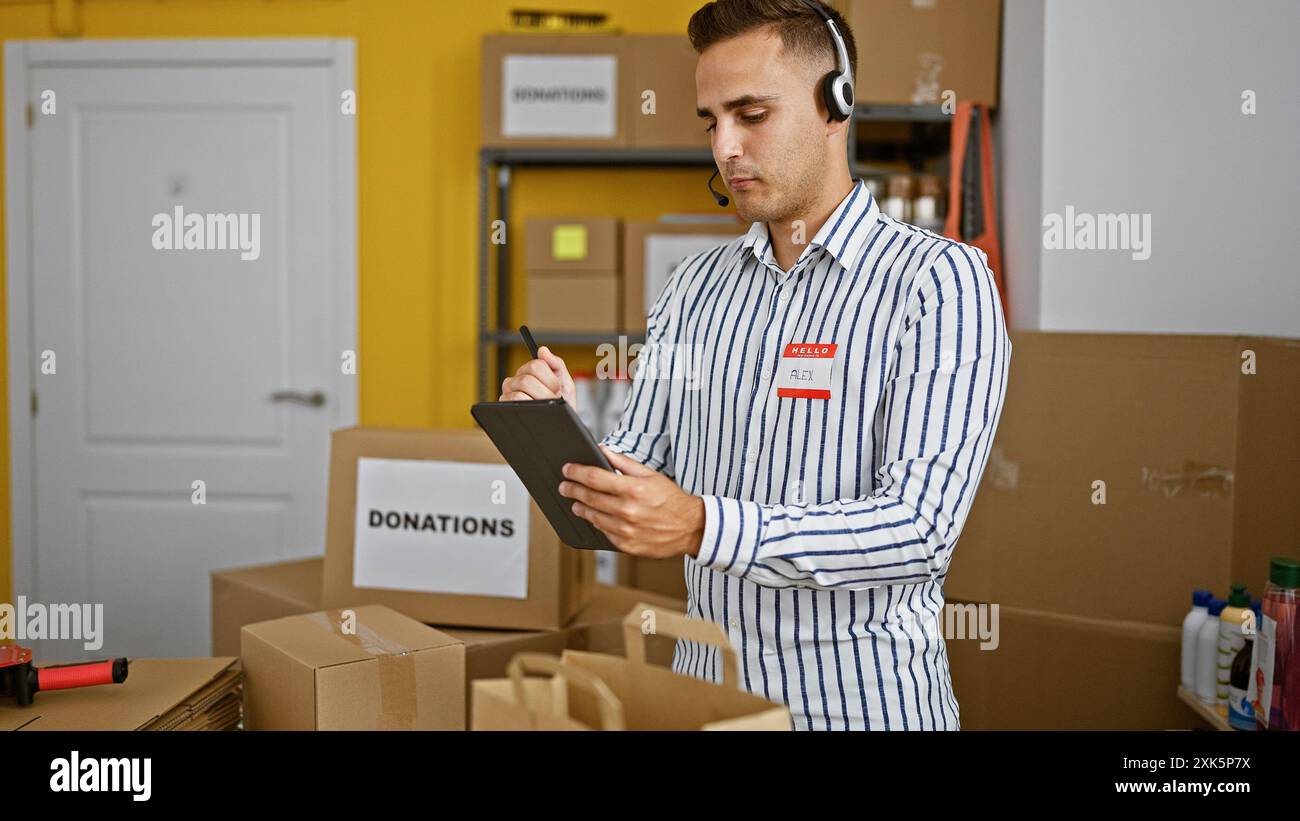 A young man wearing a headset and badge marks inventory on a tablet in a donation center warehouse. Stock Photo